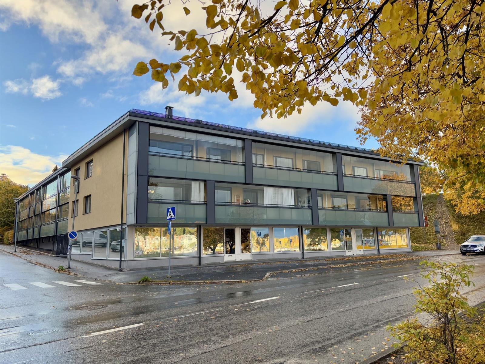 Kolmikerroksinen kerrostalo jonka alimmassa kerroksessa on liikehuoneistoja. Etualalla syksyisen lehtipuun oksa.