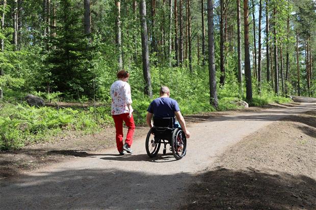 Mies ja nainen Tyysterniemen rantaraitilla. Mies istuu pyörätuolissa ja kulkee pitkin metsäreittiä nainen vierellään.